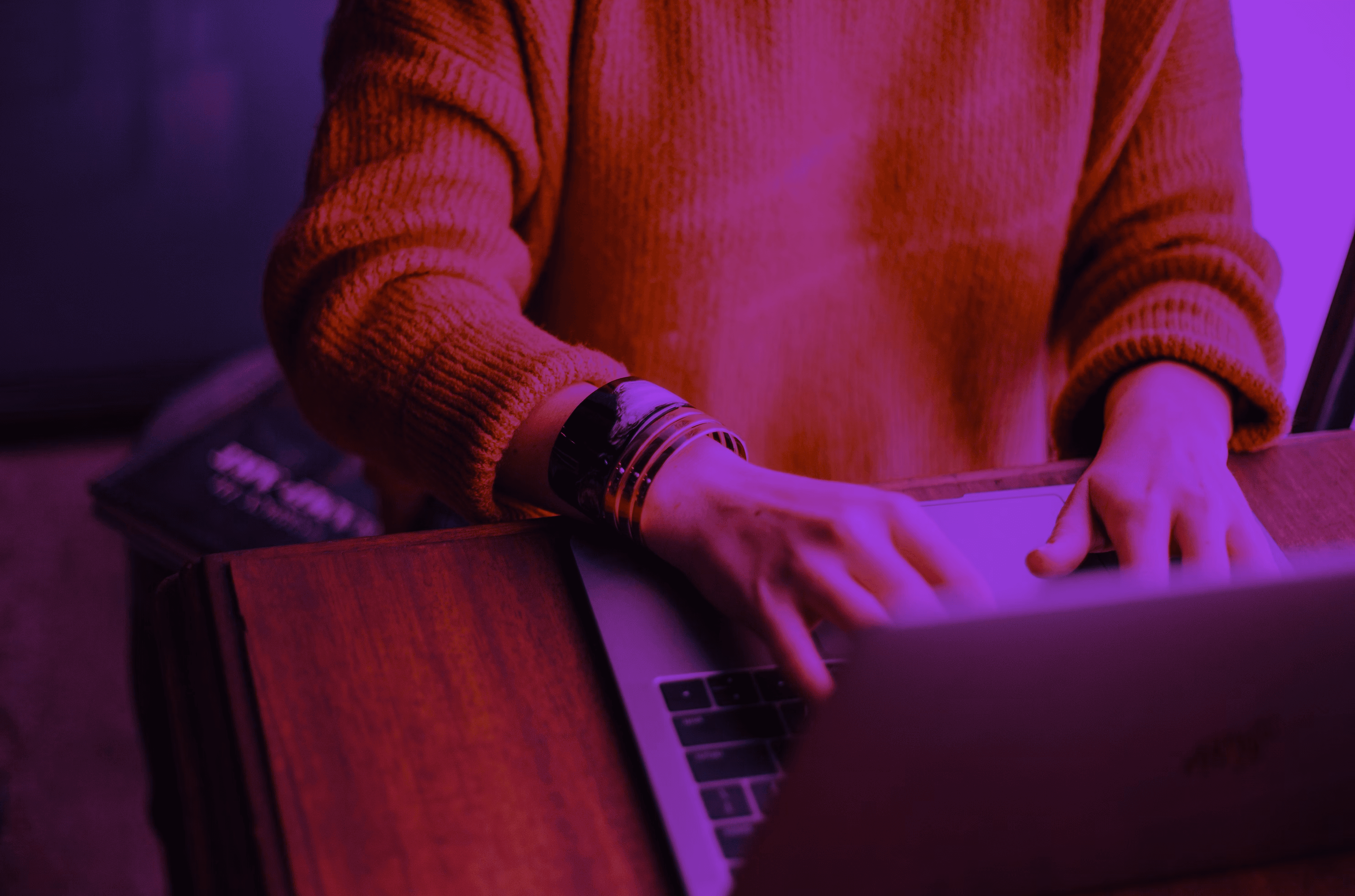 Person in a sweater typing on a laptop under dramatic pink and purple lighting.