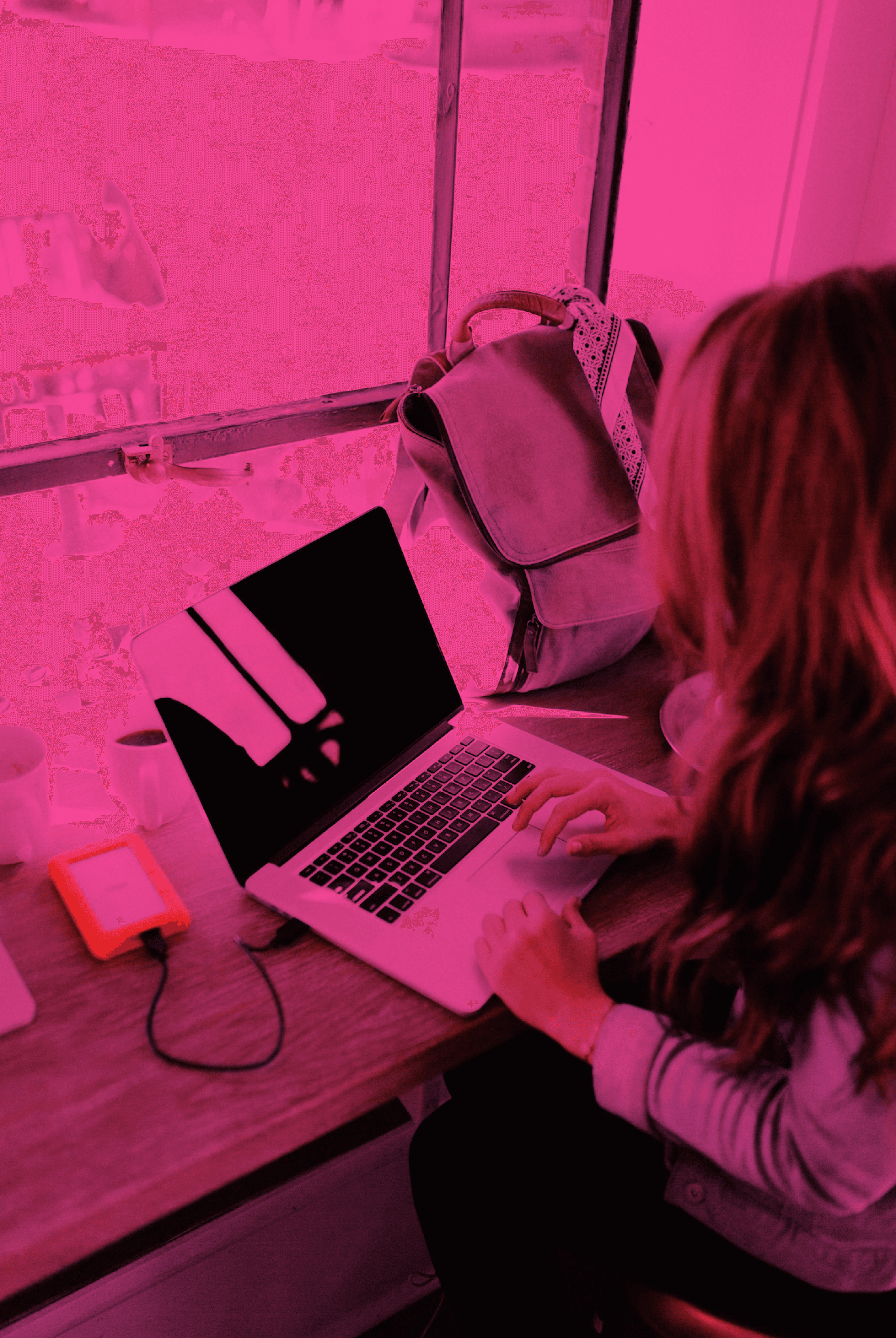 Woman working on a laptop by a window with pink filter and orange hard drive.